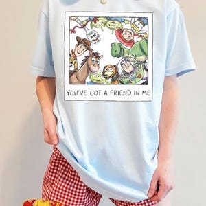 May include: Light blue t-shirt featuring a framed graphic of Toy Story characters, including Woody, Buzz Lightyear, and the aliens. The text "YOU'VE GOT A FRIEND IN ME" is below the image. The model is wearing red and white checkered shorts.