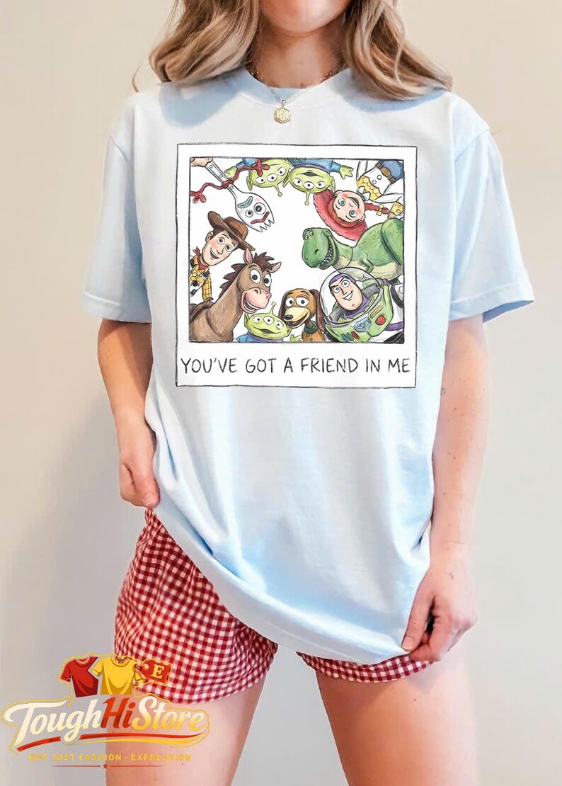 May include: Light blue t-shirt featuring a framed graphic of Toy Story characters, including Woody, Buzz Lightyear, and the aliens. The text "YOU'VE GOT A FRIEND IN ME" is below the image. The model is wearing red and white checkered shorts.