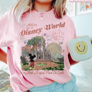 K&ouml;nnte beinhalten: Ein hellrosa T-Shirt mit einem Grafikdruck von Disney World in Florida, einschlie&szlig;lich Schloss, Epcot-Sph&auml;re und Mickey Mouse. Der Text lautet Florida Est. 1971, Disney World und The Most Magical Place On Earth in einer Vintage-Schrift.