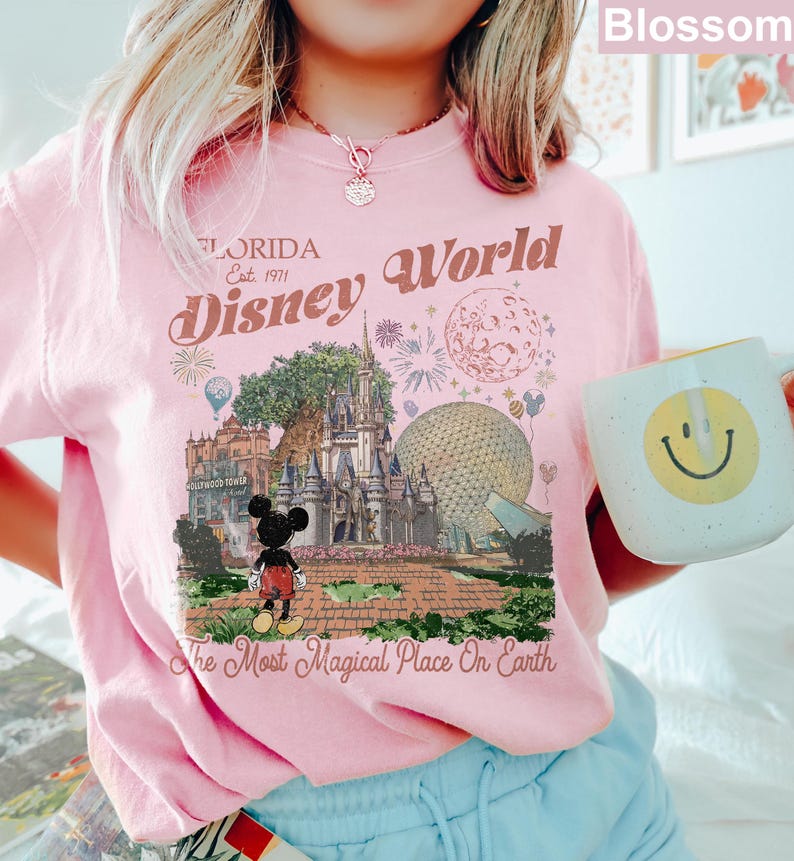 K&ouml;nnte beinhalten: Ein hellrosa T-Shirt mit einem Grafikdruck von Disney World in Florida, einschlie&szlig;lich Schloss, Epcot-Sph&auml;re und Mickey Mouse. Der Text lautet Florida Est. 1971, Disney World und The Most Magical Place On Earth in einer Vintage-Schrift.