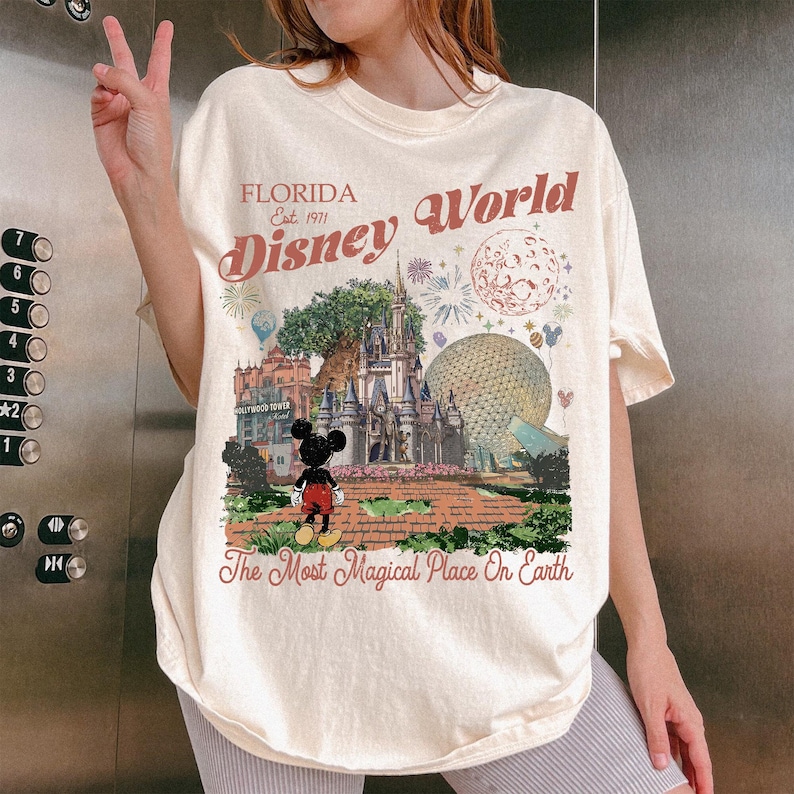 K&ouml;nnte beinhalten: Ein cremefarbenes T-Shirt mit einem Grafikdruck von Florida Disney World, einschlie&szlig;lich Schloss, Epcot-Sph&auml;re und Mickey Mouse. Der Text lautet Florida Est. 1971 Disney World und The Most Magical Place On Earth in rotbrauner Schrift.