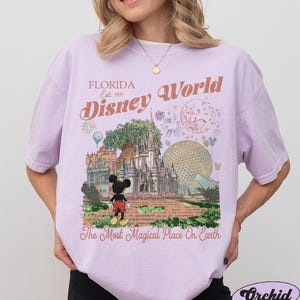 K&ouml;nnte beinhalten: Ein hellviolettes T-Shirt mit einem Grafikdruck von Disney World, auf dem Mickey Mouse vor dem Schloss steht. Der Text lautet Florida Est. 1971 Disney World und The Most Magical Place On Earth, erg&auml;nzt durch Feuerwerk und Symbole.