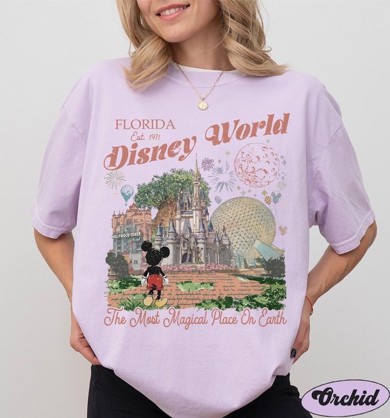 K&ouml;nnte beinhalten: Ein hellviolettes T-Shirt mit einem Grafikdruck von Disney World, auf dem Mickey Mouse vor dem Schloss steht. Der Text lautet Florida Est. 1971 Disney World und The Most Magical Place On Earth, erg&auml;nzt durch Feuerwerk und Symbole.