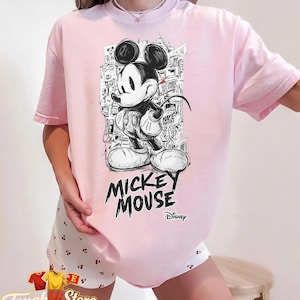 May include: A pale pink t-shirt with a black and white Mickey Mouse graphic. The design includes the words "Mickey Mouse" and the Disney logo. The shirt is from the "Tough Hill Store" collection.