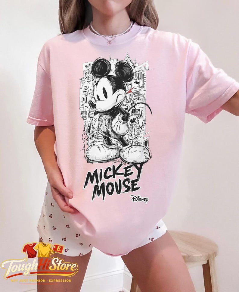 May include: A pale pink t-shirt with a black and white Mickey Mouse graphic. The design includes the words "Mickey Mouse" and the Disney logo. The shirt is from the "Tough Hill Store" collection.
