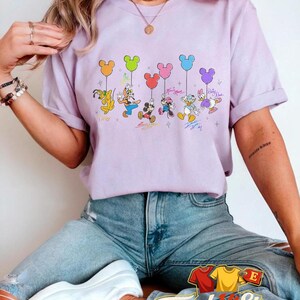 May include: A light purple t-shirt featuring a graphic of Disney characters holding Mickey Mouse-shaped balloons. The characters include Goofy, Donald Duck, Mickey Mouse, Minnie Mouse, and Daisy Duck. The shirt has the text "Tough Hit Store" at the bottom.