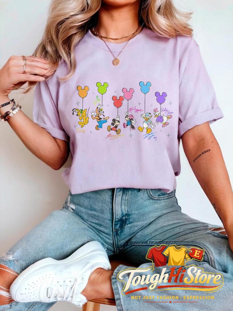 May include: A light purple t-shirt featuring a graphic of Disney characters holding Mickey Mouse-shaped balloons. The characters include Goofy, Donald Duck, Mickey Mouse, Minnie Mouse, and Daisy Duck. The shirt has the text "Tough Hit Store" at the bottom.