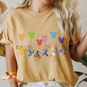 May include: Mustard yellow t-shirt featuring Disney characters holding Mickey Mouse-shaped balloons. Pluto, Goofy, Mickey, Minnie, Donald, and Daisy are depicted with their signatures below. The shirt has a vintage, distressed look.