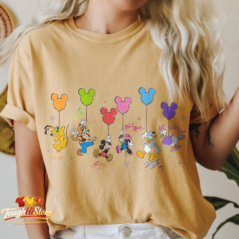 May include: Mustard yellow t-shirt featuring Disney characters holding Mickey Mouse-shaped balloons. Pluto, Goofy, Mickey, Minnie, Donald, and Daisy are depicted with their signatures below. The shirt has a vintage, distressed look.