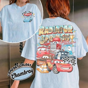 May include: Light blue t-shirt featuring a graphic of characters from the movie Cars. The back of the shirt displays "Radiator Springs" and "Lightning McQueen" with various car illustrations. The front has a small car graphic and the word "Name".
