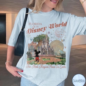 K&ouml;nnte beinhalten: Ein hellblaues T-Shirt mit einem Grafikdruck des Themenparks Disney World in Florida, einschlie&szlig;lich Schloss, Epcot-Sph&auml;re und Mickey Mouse. Der Text lautet Florida Est. 1971, Disney World und The Most Magical Place On Earth.