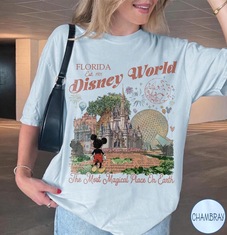 K&ouml;nnte beinhalten: Ein hellblaues T-Shirt mit einem Grafikdruck des Themenparks Disney World in Florida, einschlie&szlig;lich Schloss, Epcot-Sph&auml;re und Mickey Mouse. Der Text lautet Florida Est. 1971, Disney World und The Most Magical Place On Earth.