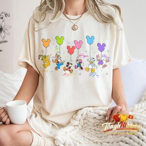 May include: Cream-colored t-shirt featuring Disney characters holding colorful Mickey Mouse-shaped balloons. The characters include Pluto, Goofy, Mickey, Minnie, Donald, and Daisy Duck. The shirt has the text "Tough Hi Store" at the bottom.