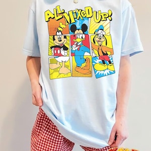 Puede incluir: Camiseta azul claro con un gr&aacute;fico colorido de Mickey Mouse, el Pato Donald y Goofy con el texto "ALL MIXED UP!". El dise&ntilde;o es de estilo c&oacute;mic. Una prenda de vestir informal.