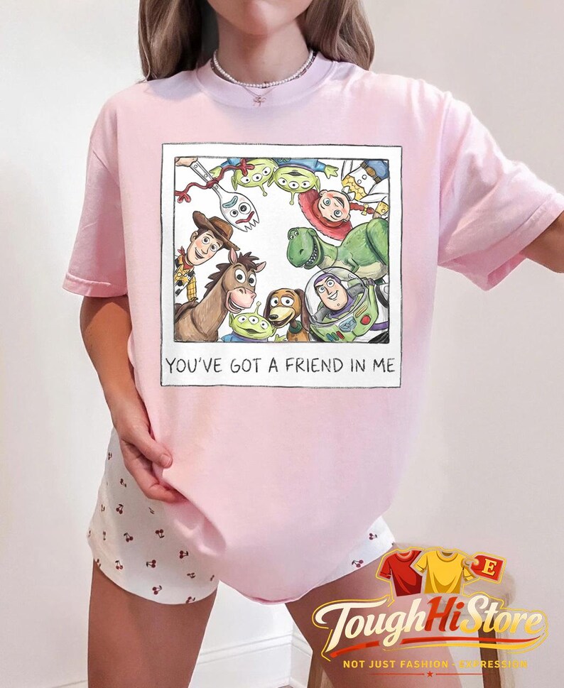 May include: A light pink t-shirt featuring a framed illustration of characters from the animated film Toy Story. The illustration includes Woody, Buzz Lightyear, and other characters. The text "YOU'VE GOT A FRIEND IN ME" is printed below the illustration.