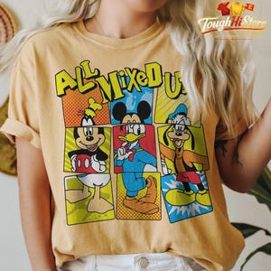 Puede incluir: Camiseta de color mostaza con un dise&ntilde;o estilo c&oacute;mic y el texto "ALL MIXED UP". El dise&ntilde;o incluye a Mickey Mouse, el Pato Donald y Goofy. La camiseta tiene mangas cortas y un corte holgado.