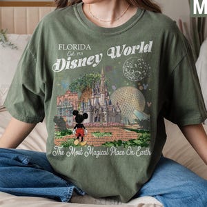 K&ouml;nnte beinhalten: Ein moosgr&uuml;nes T-Shirt mit einem Grafikdruck des Disney World Schlosses, der Epcot-Kugel und Mickey Mouse. Der Text lautet Florida Est. 1971 Disney World und The Most Magical Place On Earth. Das Design enth&auml;lt einen Mond und Sterne.
