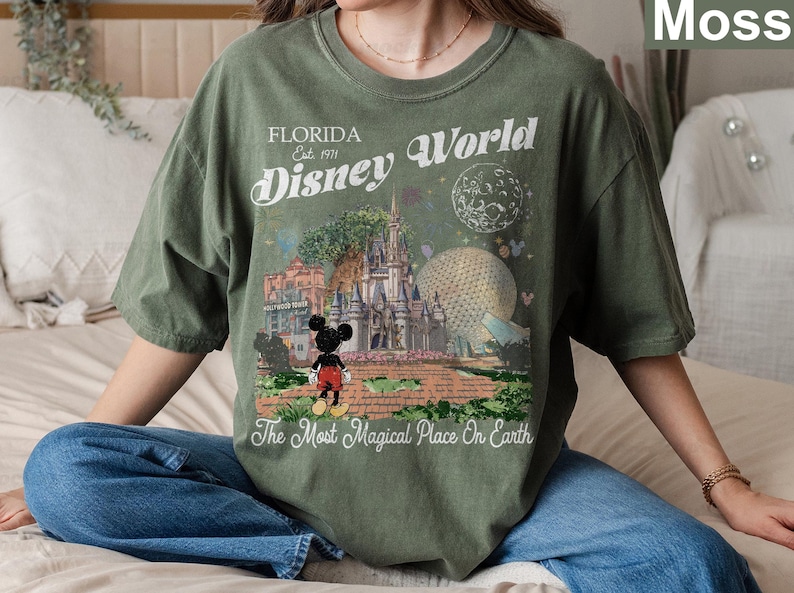 K&ouml;nnte beinhalten: Ein moosgr&uuml;nes T-Shirt mit einem Grafikdruck des Disney World Schlosses, der Epcot-Kugel und Mickey Mouse. Der Text lautet Florida Est. 1971 Disney World und The Most Magical Place On Earth. Das Design enth&auml;lt einen Mond und Sterne.