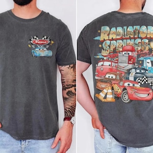 May include: Charcoal gray t-shirt featuring a cartoon car design on the front with the word "Name" above it. The back of the shirt displays a colorful graphic of various cartoon cars with the text "Radiator Springs".