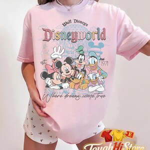 Puede incluir: Camiseta rosa claro con un dise&ntilde;o de Disney que presenta a Mickey y Minnie Mouse, Goofy, Donald y Daisy Duck. La camiseta tiene el texto "Walt Disney's Disneyworld" y "Where dreams come true".