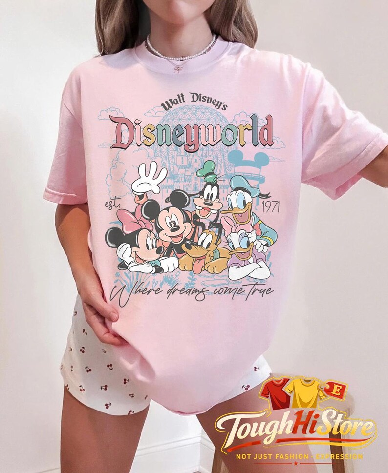 Puede incluir: Camiseta rosa claro con un dise&ntilde;o de Disney que presenta a Mickey y Minnie Mouse, Goofy, Donald y Daisy Duck. La camiseta tiene el texto "Walt Disney's Disneyworld" y "Where dreams come true".