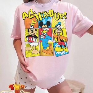 Puede incluir: Camiseta rosa claro con un dise&ntilde;o de c&oacute;mic de Mickey, Donald y Goofy, con el texto "ALL MIXED UP!". El dise&ntilde;o utiliza colores brillantes y contornos. Parte de una colecci&oacute;n de ropa.