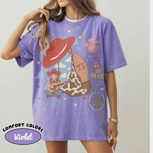 May include: A violet short-sleeve t-shirt featuring a graphic print of a red cowboy hat, a cow-print boot with the initials TS, a cartoon character playing a guitar, a pink disco ball hat, alphabet blocks, and a cactus surrounded by musical notes.