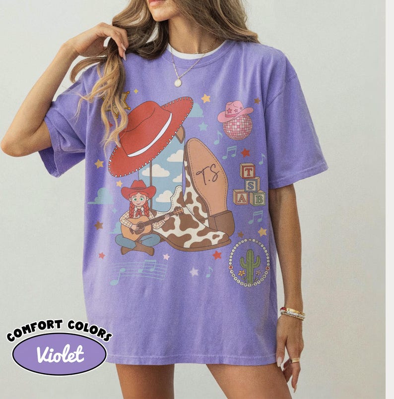 May include: A violet short-sleeve t-shirt featuring a graphic print of a red cowboy hat, a cow-print boot with the initials TS, a cartoon character playing a guitar, a pink disco ball hat, alphabet blocks, and a cactus surrounded by musical notes.