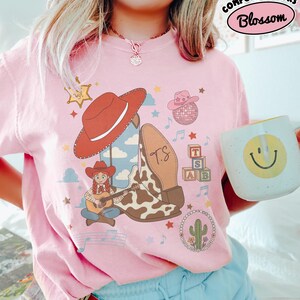 May include: A light pink t-shirt featuring a graphic print of a red cowboy hat, a cow-print boot with the letters TS, a cartoon character playing a guitar, a disco ball, alphabet blocks, a cactus, and musical notes. The text Comfort Colors Blossom is visible.