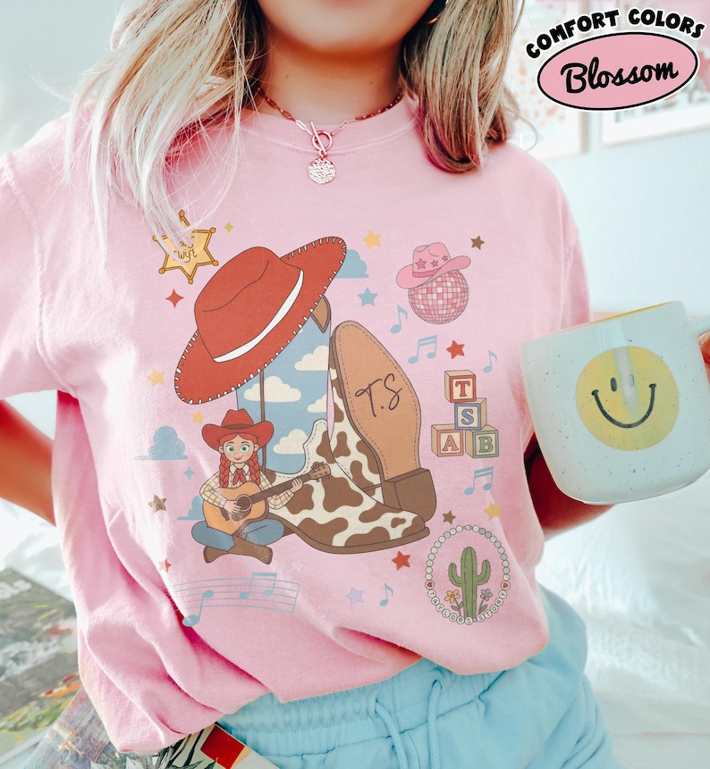 May include: A light pink t-shirt featuring a graphic print of a red cowboy hat, a cow-print boot with the letters TS, a cartoon character playing a guitar, a disco ball, alphabet blocks, a cactus, and musical notes. The text Comfort Colors Blossom is visible.