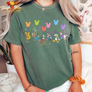 May include: Green t-shirt featuring Disney characters holding Mickey Mouse-shaped balloons. Pluto, Goofy, Mickey, Minnie, Donald, and Daisy are depicted. The shirt has the text "Tough It Store" at the top.