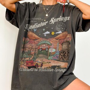 K&ouml;nnte beinhalten: Ein anthrazitfarbenes Kurzarm-T-Shirt mit einem Grafikdruck von Radiator Springs. Das Design zeigt rote Rennwagen, W&uuml;stenfelsformationen, einen Wasserturm sowie den Text California Est 2012, Radiator Springs und Welcome to Radiator Springs.