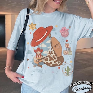 May include: A light blue t-shirt featuring a graphic print of a red cowboy hat, a cow-print boot with the initials T.S., a cartoon character playing a guitar, a sheriff star, a disco ball, alphabet blocks, and a cactus inside a beaded circle.