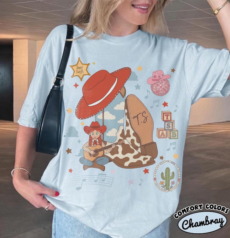 May include: A light blue t-shirt featuring a graphic print of a red cowboy hat, a cow-print boot with the initials T.S., a cartoon character playing a guitar, a sheriff star, a disco ball, alphabet blocks, and a cactus inside a beaded circle.