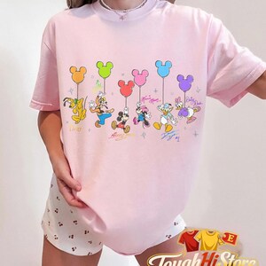 May include: A light pink t-shirt featuring a graphic of Disney characters holding Mickey Mouse-shaped balloons. The characters include Pluto, Goofy, Mickey, Minnie, Donald, and Daisy. Each character has a signature below them. The shirt has short sleeves.