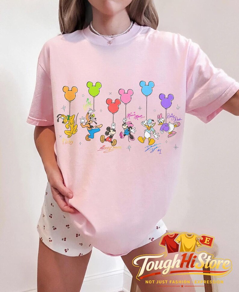 May include: A light pink t-shirt featuring a graphic of Disney characters holding Mickey Mouse-shaped balloons. The characters include Pluto, Goofy, Mickey, Minnie, Donald, and Daisy. Each character has a signature below them. The shirt has short sleeves.