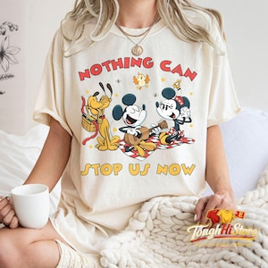 May include: Cream-colored t-shirt featuring Mickey Mouse, Minnie Mouse, and Pluto having a picnic. The graphic includes the text "NOTHING CAN STOP US NOW" in red and yellow. The shirt is a relaxed fit.