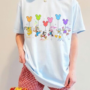 May include: Light blue t-shirt featuring Disney characters holding Mickey Mouse-shaped balloons in various colors. The characters include Pluto, Goofy, Mickey Mouse, Minnie Mouse, Donald Duck, and Daisy Duck. Signatures are below each character.