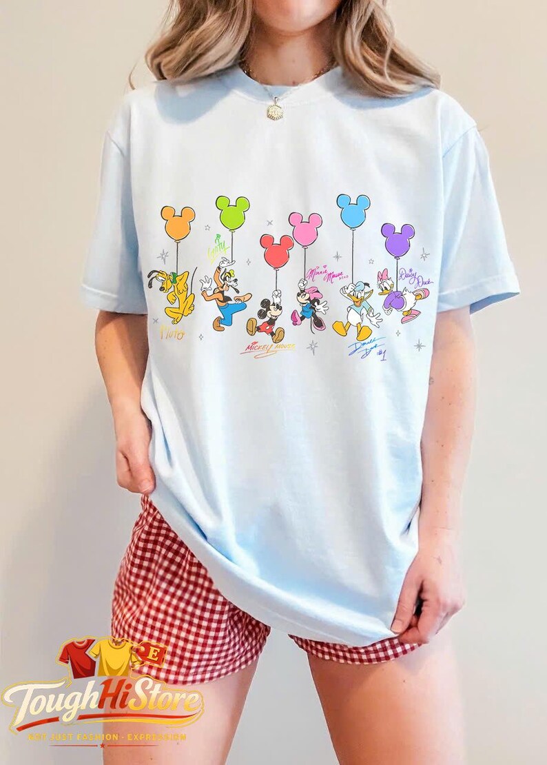 May include: Light blue t-shirt featuring Disney characters holding Mickey Mouse-shaped balloons in various colors. The characters include Pluto, Goofy, Mickey Mouse, Minnie Mouse, Donald Duck, and Daisy Duck. Signatures are below each character.
