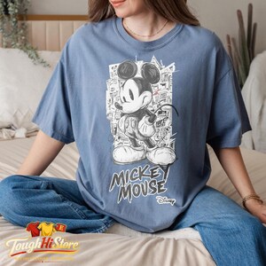 May include: A blue t-shirt with a black and white Mickey Mouse illustration. The design includes the words "Mickey Mouse" in a stylized font, with the Disney logo underneath.