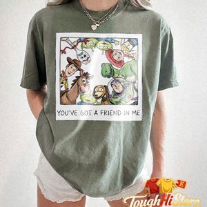May include: Olive green t-shirt featuring a framed illustration of characters from the movie Toy Story. The illustration includes Woody, Buzz Lightyear, and other characters. The text "YOU'VE GOT A FRIEND IN ME" is below the illustration.