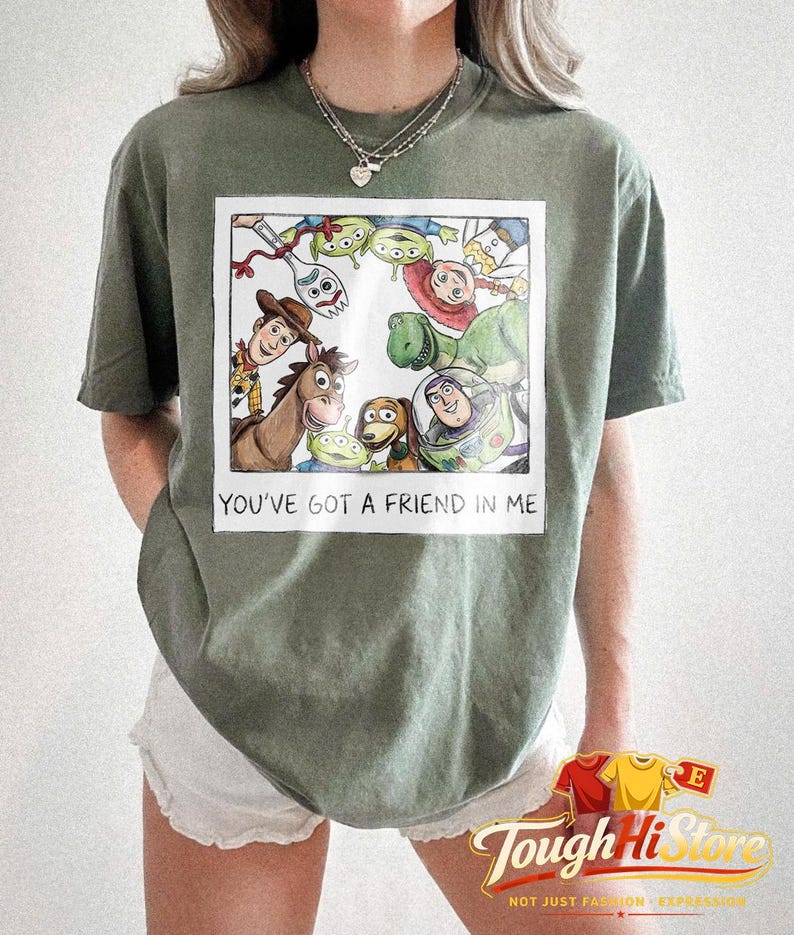 May include: Olive green t-shirt featuring a framed illustration of characters from the movie Toy Story. The illustration includes Woody, Buzz Lightyear, and other characters. The text "YOU'VE GOT A FRIEND IN ME" is below the illustration.