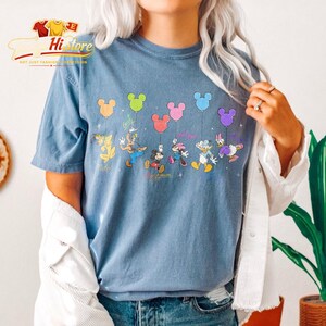 May include: A blue t-shirt featuring a colorful graphic of Disney characters holding Mickey Mouse-shaped balloons. The characters include Pluto, Goofy, Mickey Mouse, Minnie Mouse, Daisy Duck, and Donald Duck. The shirt has the text "Walt Disney World" and "Est. 1971".