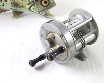 Puede incluir: Un carrete de pesca vintage con acabado plateado y mangos negros. El carrete está colocado frente a una figura de pez de cerámica con detalles moteados verdes y negros.