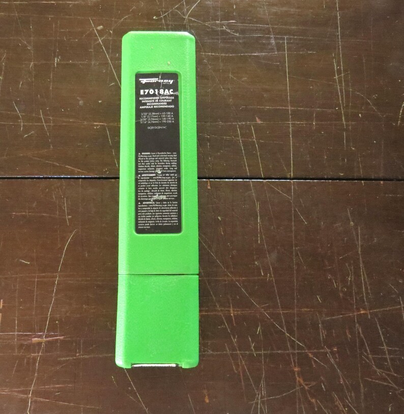 May include: A vivid green rectangular case with a black label. The label displays the text "E7018AC" and technical details. The case is placed on a dark wooden surface.