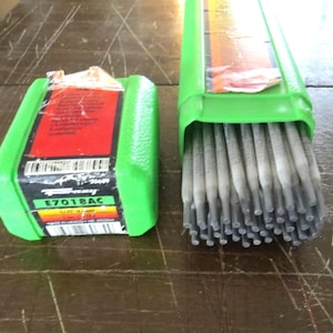 May include: Two green containers of welding rods. One is closed, the other open, showing the rods. The closed container has a red and black label with the text "E7018AC". The rods are grey.