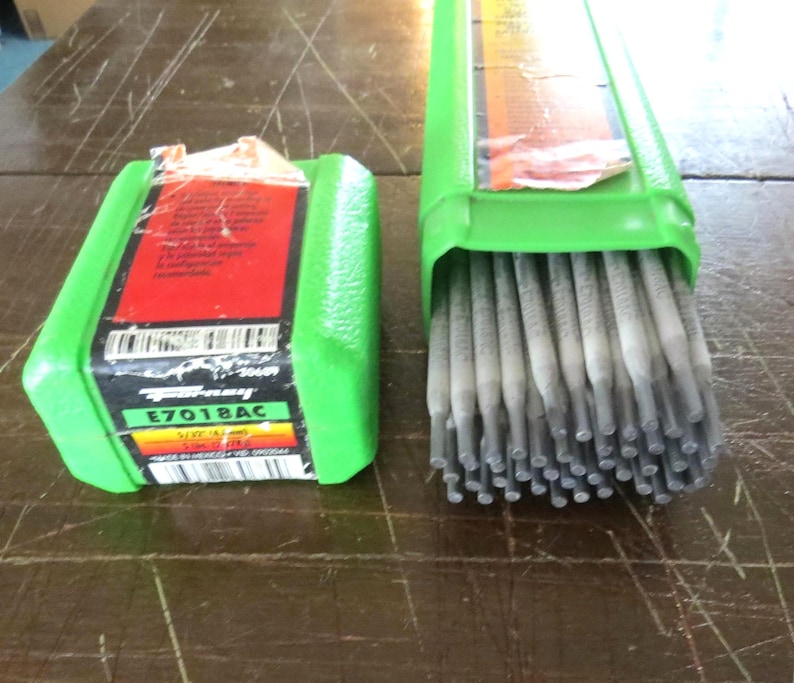 May include: Two green containers of welding rods. One is closed, the other open, showing the rods. The closed container has a red and black label with the text "E7018AC". The rods are grey.