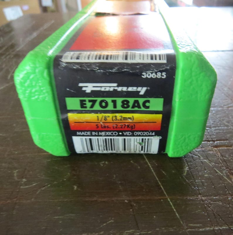 Puede incluir: Un paquete de varillas de soldadura Forney E7018AC. La etiqueta incluye el texto "1/8" (3,2 mm)" y "5 lbs. (2,27 kg)". El paquete tiene una cubierta de pl&aacute;stico verde y una etiqueta negra y roja.