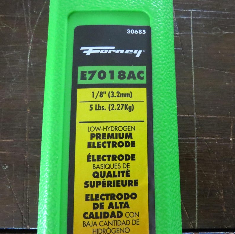 Puede incluir: Un paquete verde con la marca "Forney" y la informaci&oacute;n del producto. El texto incluye "E7018AC", "1/8" (3,2 mm)" y "5 Lbs. (2,27 kg)". El texto adicional describe el producto como un electrodo premium de bajo hidr&oacute;geno.