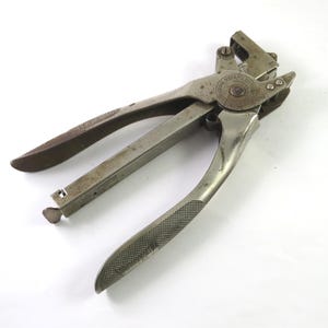May include: An antique metal tool with a hinged design, featuring textured handles and a circular emblem. The tool has a silver finish with signs of wear, suggesting age and use. The tool is designed for a specific function, likely related to fastening or shaping materials.